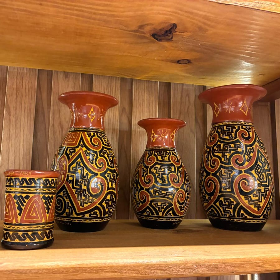 Três vasos e um copo de cerâmica com grafismo marajoara em preto, amarelo e vermelho, numa prateleira de madeira